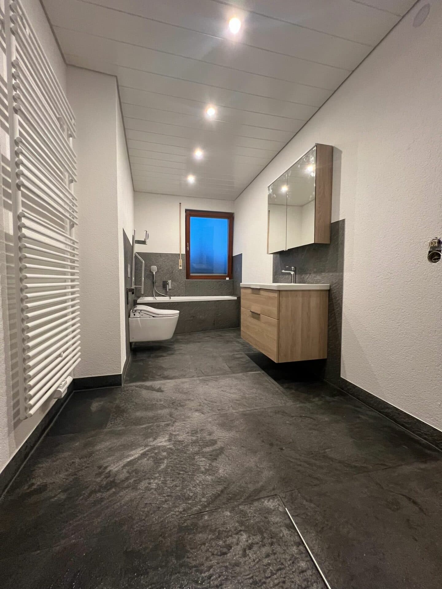 Schmales Bad mit dunkler Steinwand, Eiche-Waschtisch und eingebauter Wanne